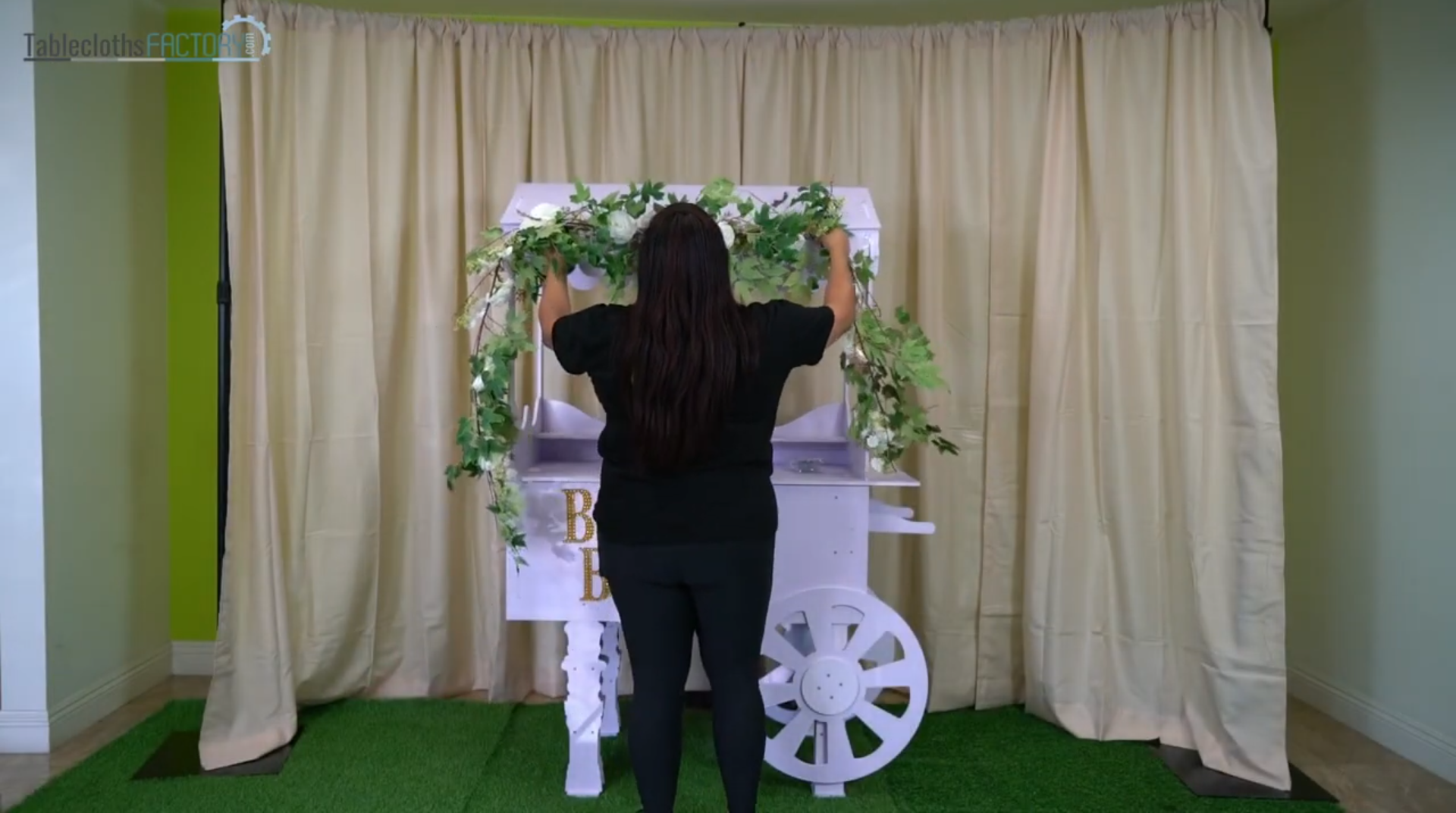 A woman arranges vines on a white cart for Mother's Day decor in a staged area.
