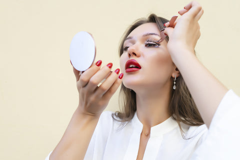 woman with short brown hair and red lipstick holding a mirror and applying false lashes. Cartel lash