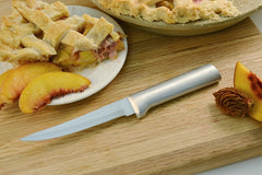 A RADA Super Parer knife next to a slice of peach pie with crisscross crust and a sliced peach to the right of the knife with a whole pie above it
