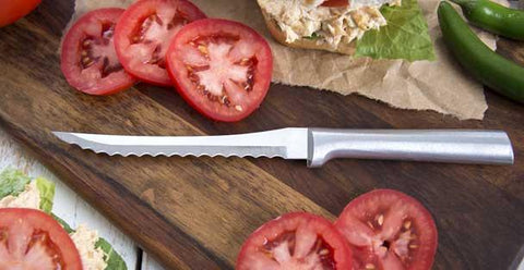 A Rada Tomato Slicer knife on a wooden cutting board next to several red slices of tomatoes and a tuna sandwich on wrapping paper