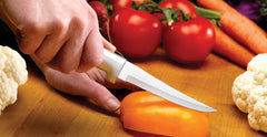 A RADA stainless steel Super Parer slicing a yellow pepper with another hand to help guide the blade with a head of cauliflower to the right a couple red tomatoes behind the Super Parer