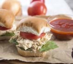A delicious pulled chicken slider in front of a few more sandwiches, a bowl of salsa and two tomatoes