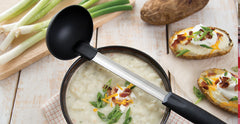 A bowl of potato soup with a RADA Non-Scratch Ladle laid over the soup with several sprigs of green onion and loaded potato halves beside the soup bowl