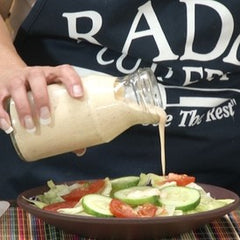 A jar of creamy cucumber onion dill dressing being poured over a salad consisting of sliced cucumbers, halved tomatoes and lettuce