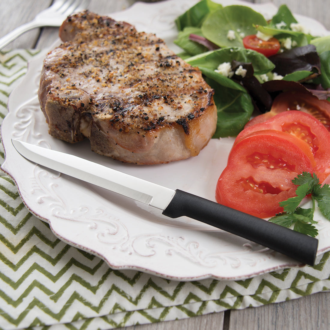 Rada Cutlery Utility Steak knife with black handle on plate with steak, spinach and tomatoes. 