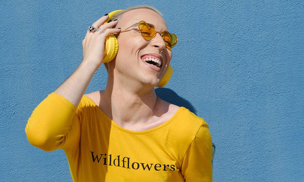 A joyful non-binary individual with a feminine male expression, wearing a yellow shirt and headphones against a yellow background, celebrating their coming out journey with happiness.