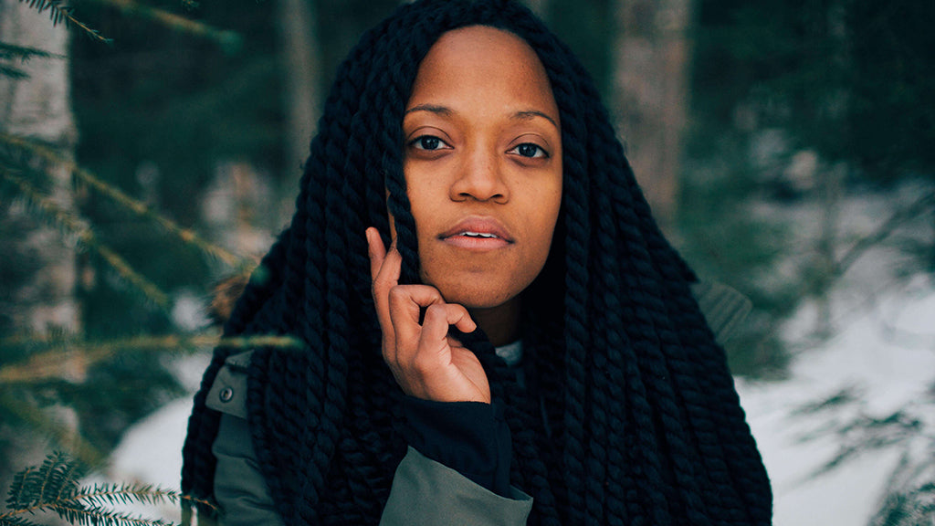 A black woman with braids stares down the lens surrounded by ferns and snow
