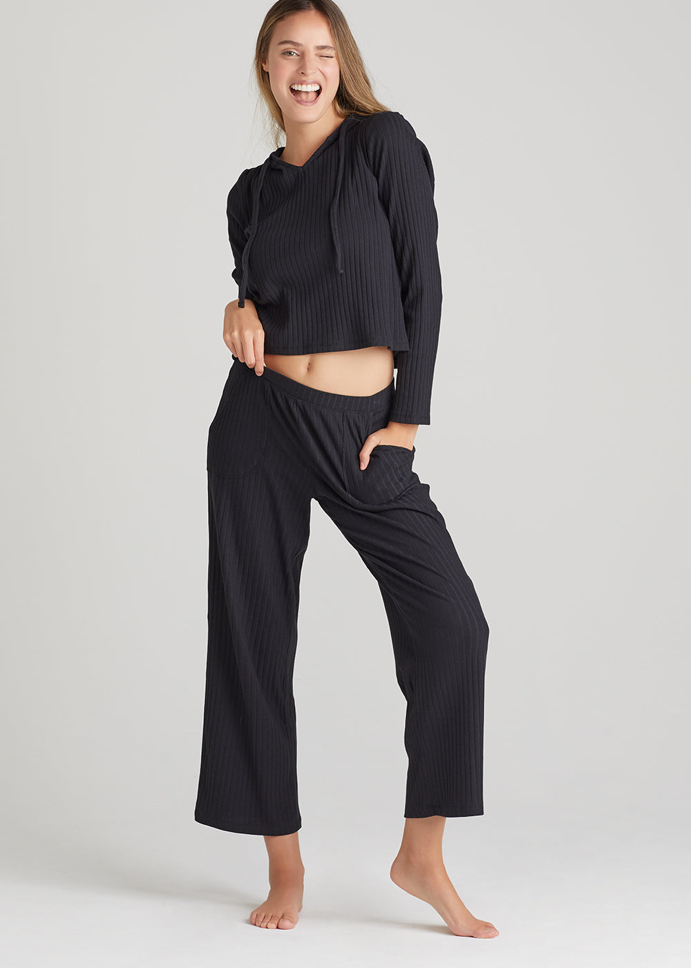 cropped lounge pant - cotton rib and cropped lounge hoodie - cotton rib in Black worn by a woman standing facing forward with right hand on hip and left hand in pocket Yummie