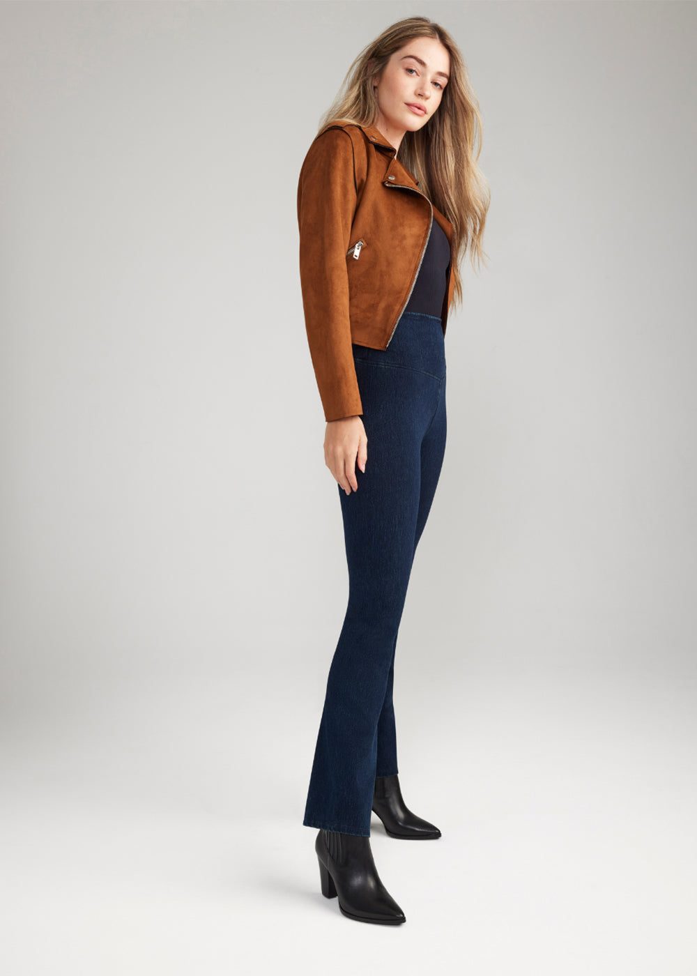 denim bootcut shaping legging in True Indigo, Black top and Suede jacket in Dark Camel worn by a woman standing sideways with arm at side Yummie