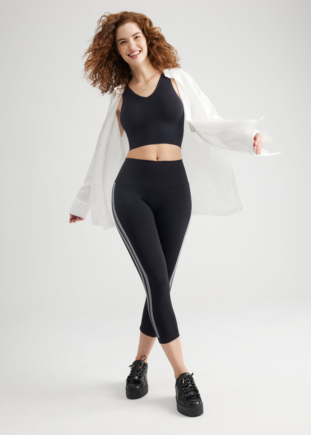 morgan seamless cropped capri shaping legging in black with white stripes standing with her right leg crossed ahead of her left with her right arm swinging forward and left arm behind Yummie