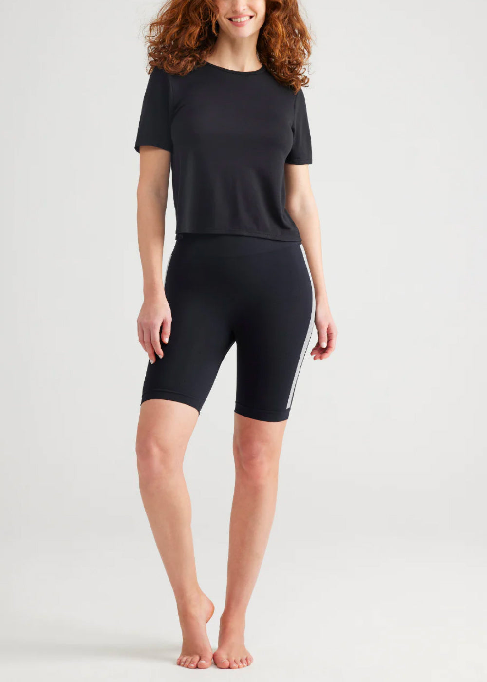 cora seamless shaping biker short in black with white stripe worn by a woman standing with her right foot out front of her and right hand on thigh Yummie