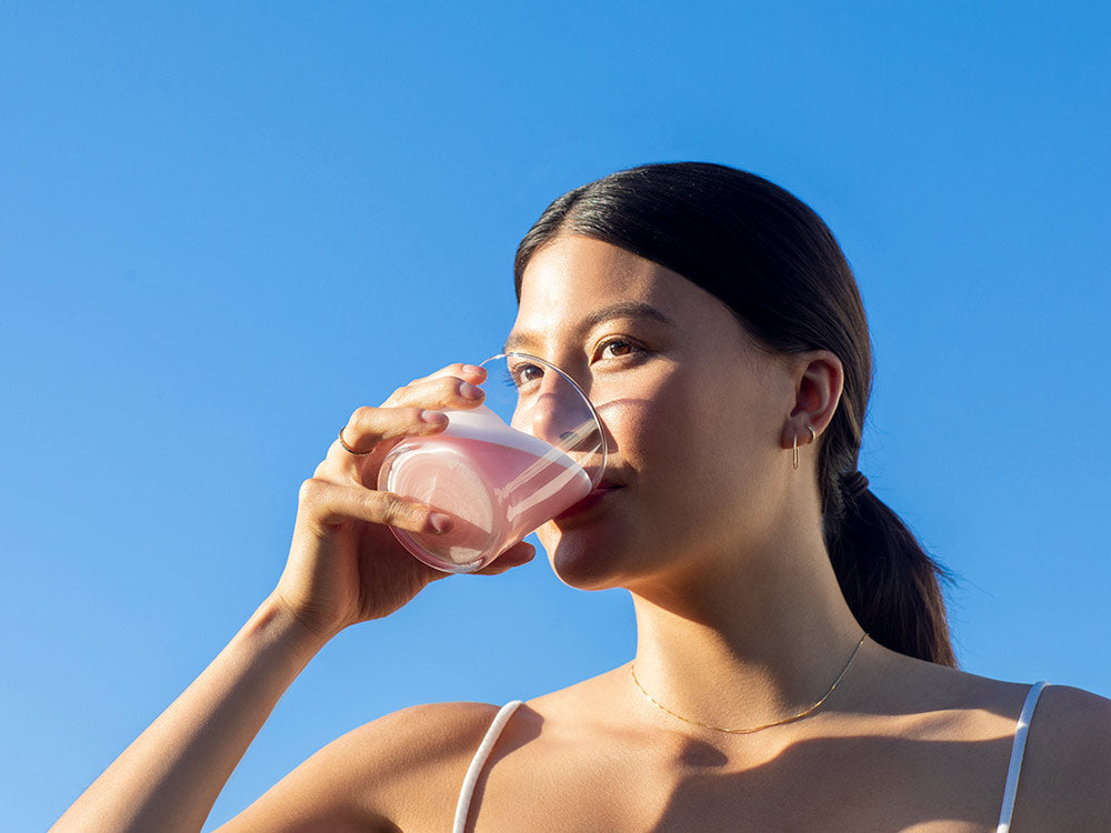 A woman drinking Vejo's Collagen Glow with the sun hitting her skin and glowing with a comforting blue sky background behind her.