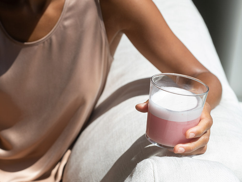 A clear glass with Vejo's Collagen Glow pink smoothie sitting in it glistening from the shine of the sun on the edge of a white sofa.