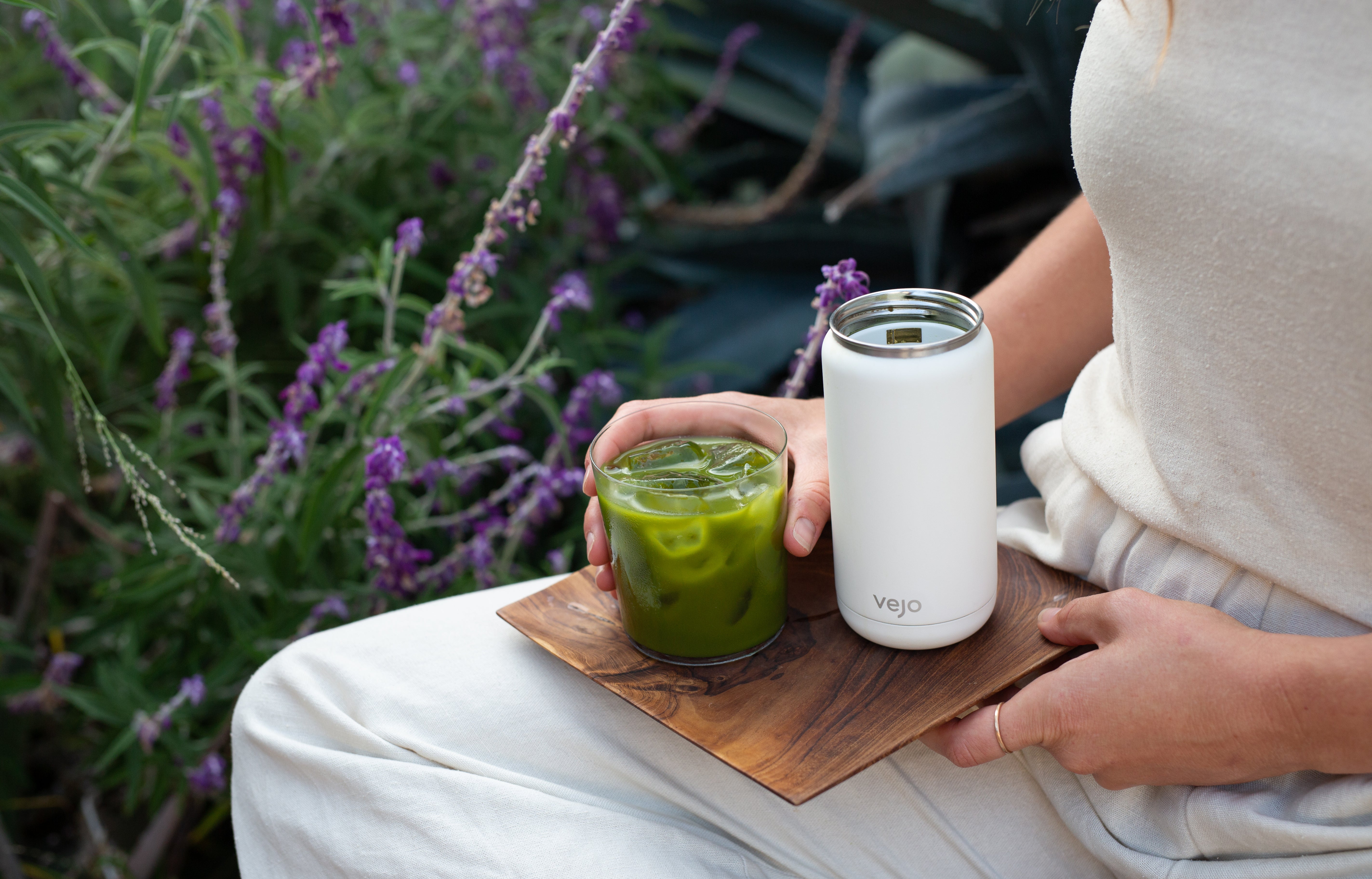 A woman holding her white Vejo and glass of Matcha next to a small grove of deep purple lavender in the morning sun.
