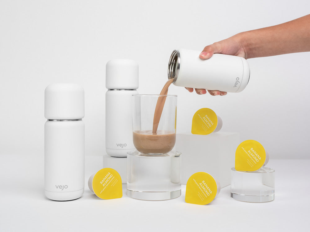 Vejo’s Banana Almond blend being poured into a clear glass by a white Vejo blender, surrounded by 2 other white Vejo blenders & Banana almond blends.