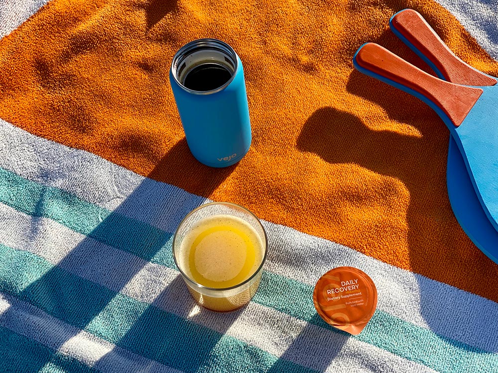 A blue vejo blender sitting on a orange and blue striped beach towel by a glass of daily recovery blend and ping pong paddles.