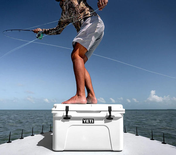 a man standing on top of a closed white YETI Tundra® 65 Cool Box  on a boat while casting his fishing line