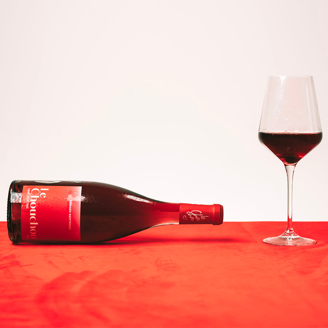 A bottle of red wine and a glass filled with wine on a red surface.