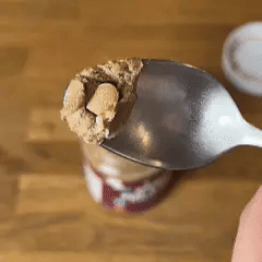 A spoon of brown powder with tiny marshmallows above a jar on a wooden surface.