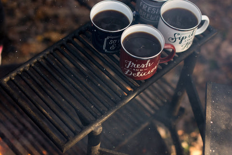 Mugs of coffee beside a campfire in the mist of making a camping cocktail.