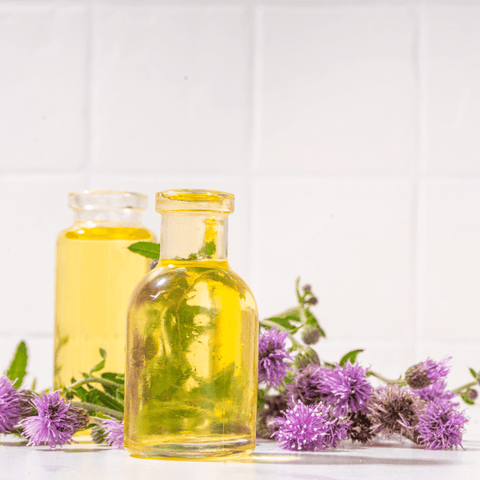 Mariendistelöl in durchsichtigen Flaschen auf einem weißen Untergrund. Als Dekoration liegen Blüten der Mariendistel daneben.