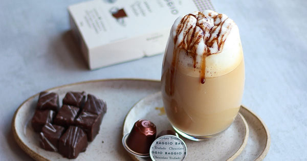 Copo de café com chocolate cremoso servido ao lado de doces de chocolate e cápsulas de café Baggio.