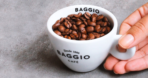 Uma mão segurando a alça de uma xícara com grãos de café no seu interior. A xícara está apoiada em uma mesa.