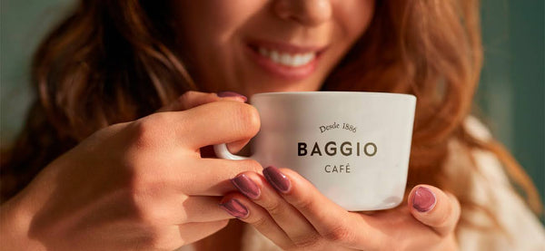 Pessoa segurando com as duas mãos uma xícara de café Baggio próxima ao rosto, pronta para beber.