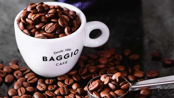 Xícara branca da Baggio repleta de grãos de café, alguns espalhados pela mesa, com um medidor ao lado.