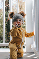 Baby kneeling on porch wearing an organic cotton footie pajama in honey with a grey knit cap that has honey colored pompoms on top.