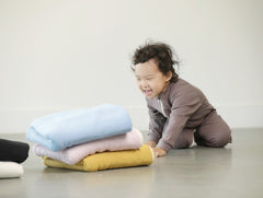 Toddler girl kneeling on floor while pushing away a stack of blankets. 