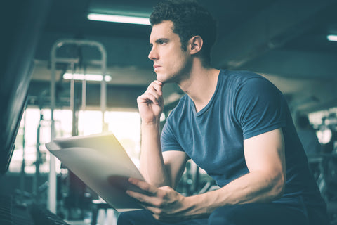Muscular handsome trainer looking at fitness plan on clipboard for working out in the fitness gym