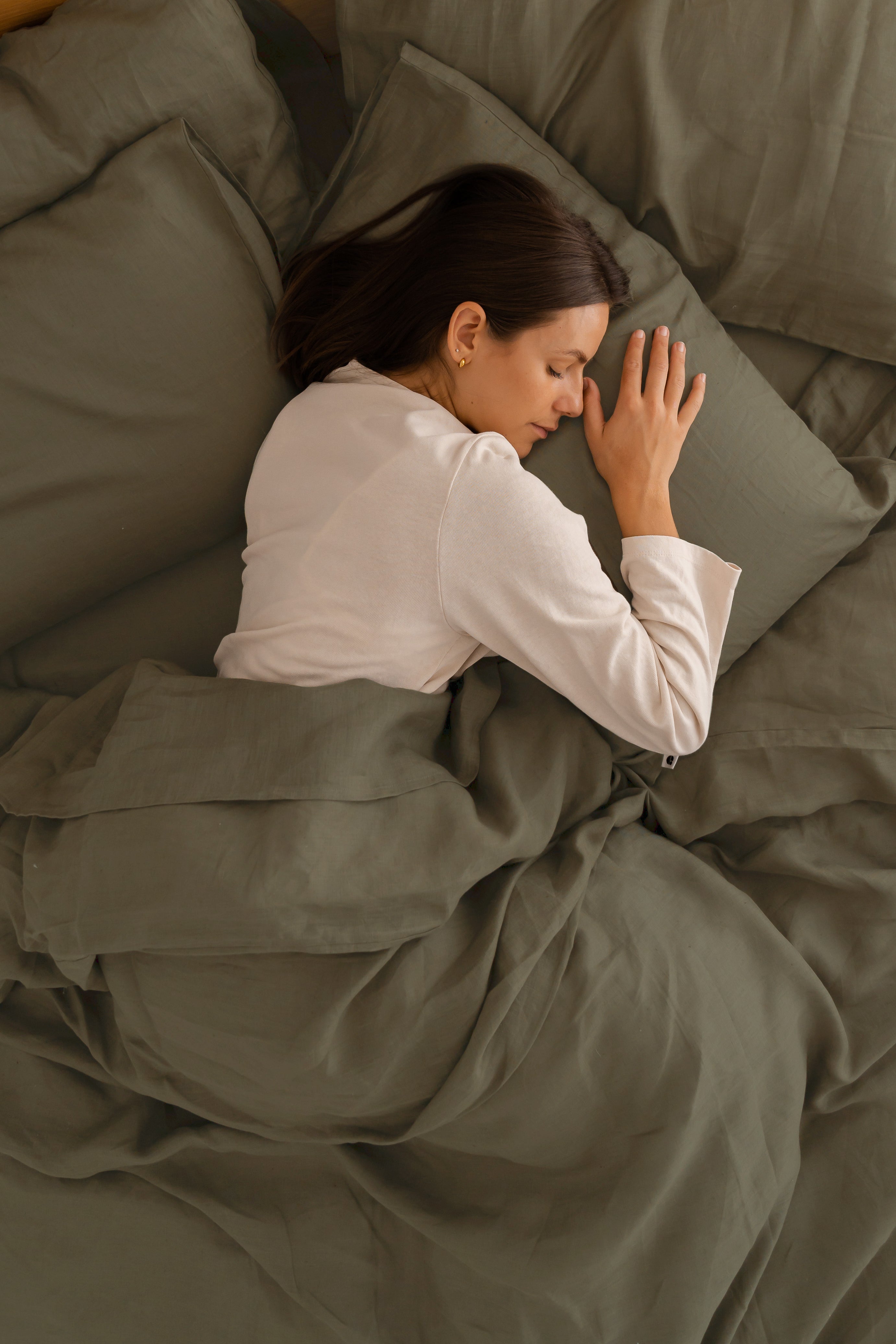 woman lying in bed, tucked in with Eva hemp linen sheets in olive