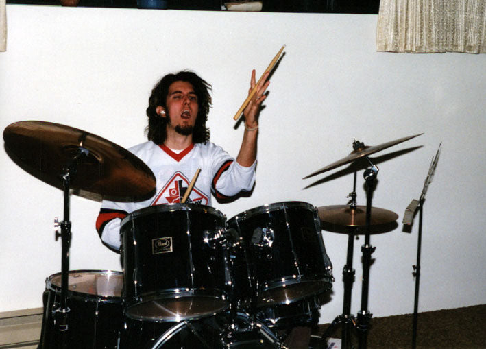 man with white t-shirt and shoulder length hair wearing a chin beard playing drums twirling a drumstick in his left hand