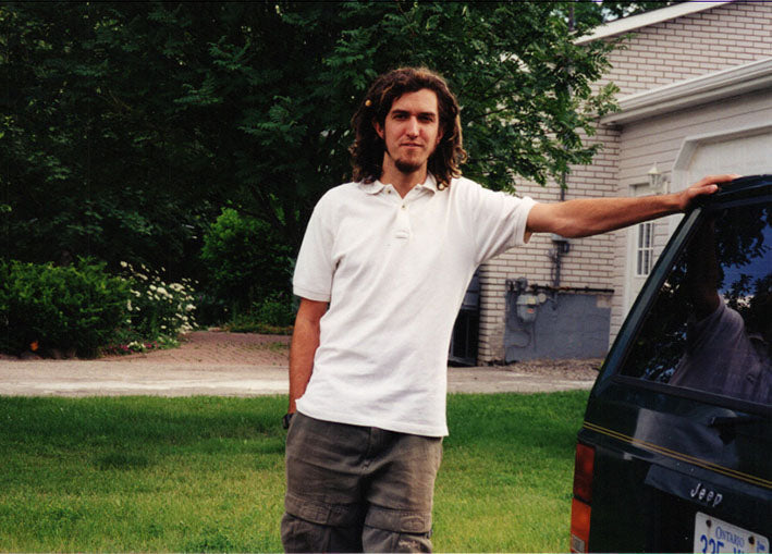 man with shoulder length dreadlocks in white t shirt leaning on minivan with green grassy lawn behind him