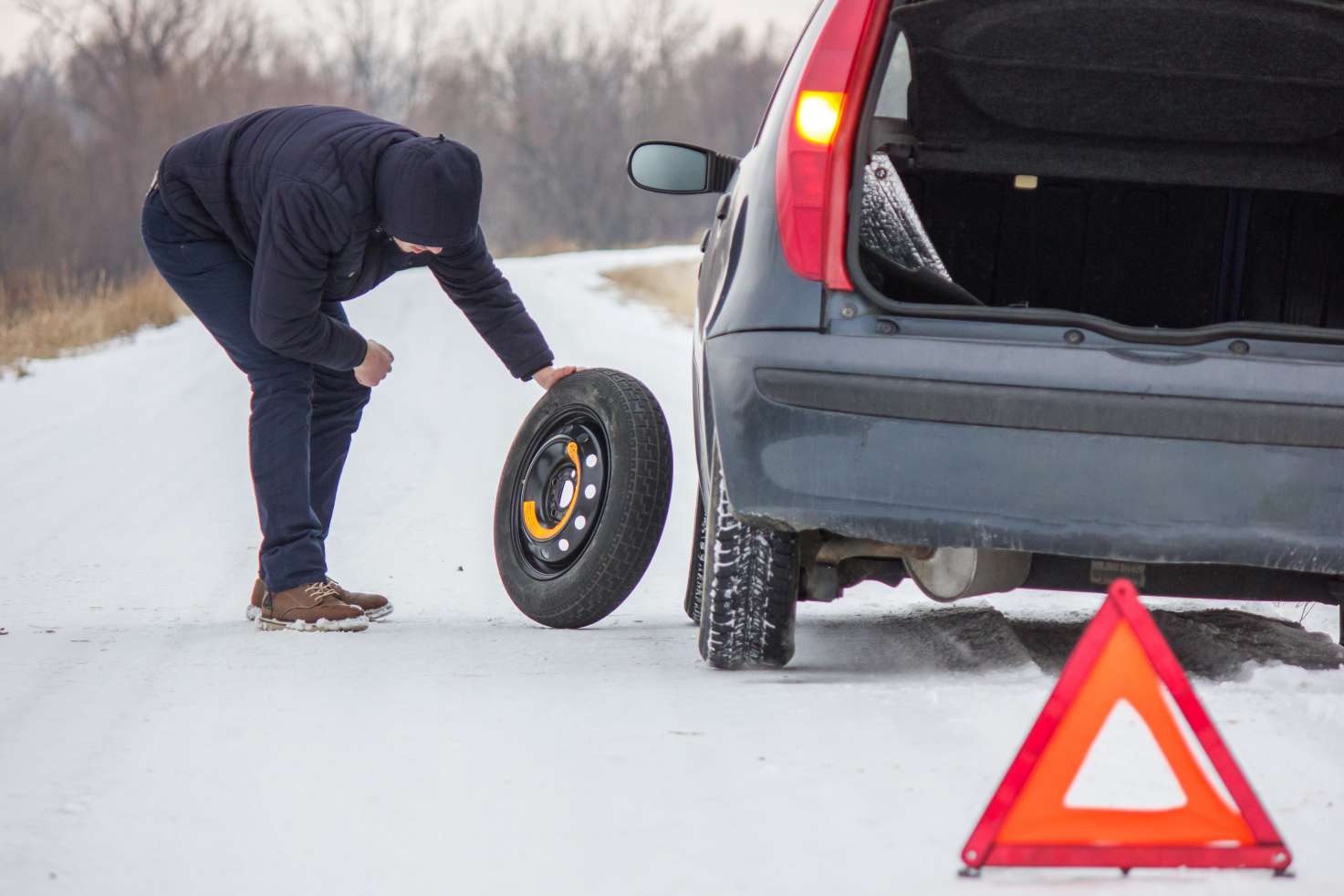 Man changing a tire on his car in the winter. A pylon is set up for passing cars.
