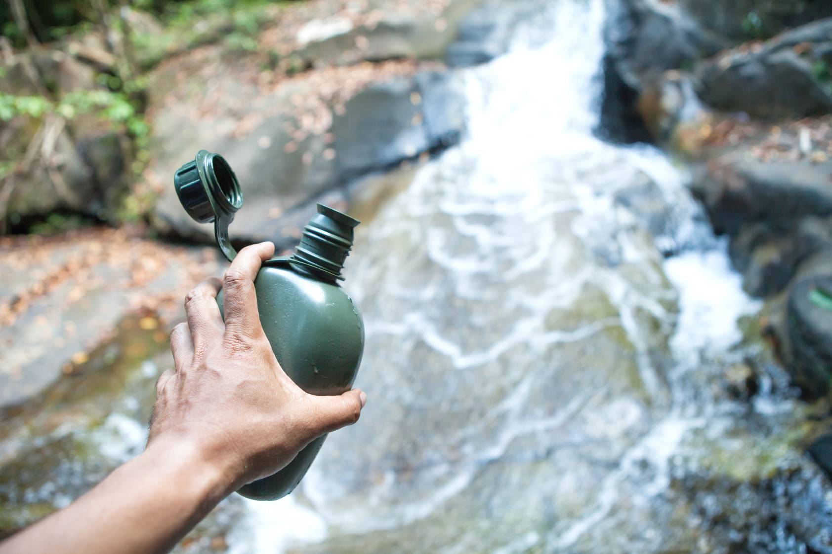 A man looking to fill up his green coloured canteen in a flowing stream of river water