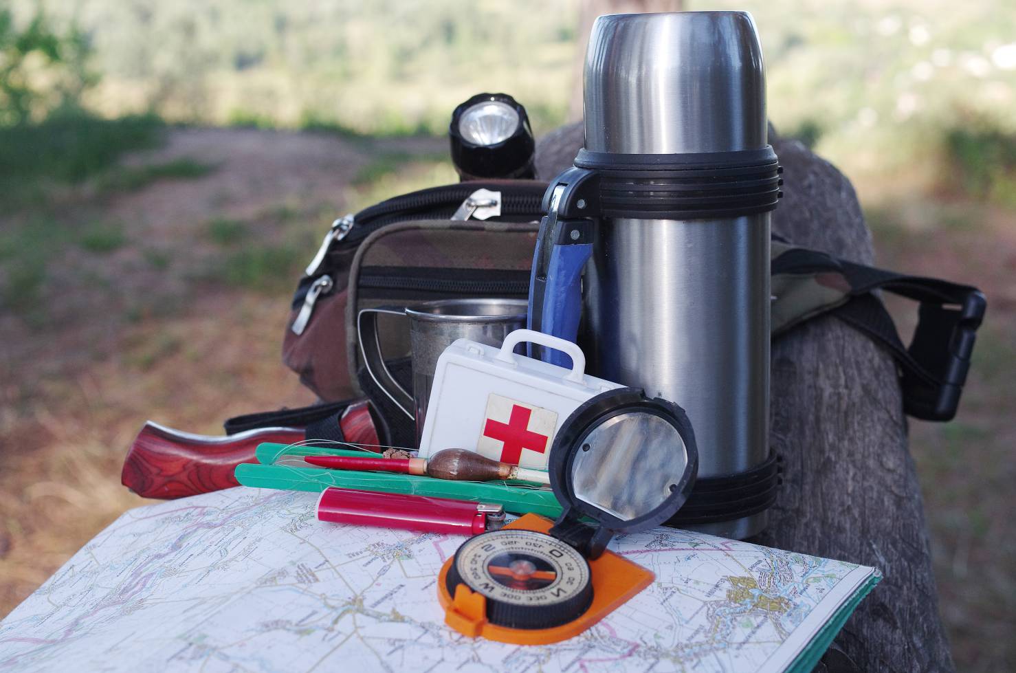 A compass, thermos, medical kit, flashlight, and knife on top of a map in the woods.