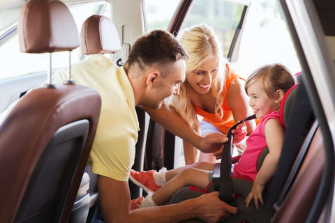 A Mother wearing a white shirt and purse and a Father wearing a yellow polo shirt buckling their infant child into a car seat