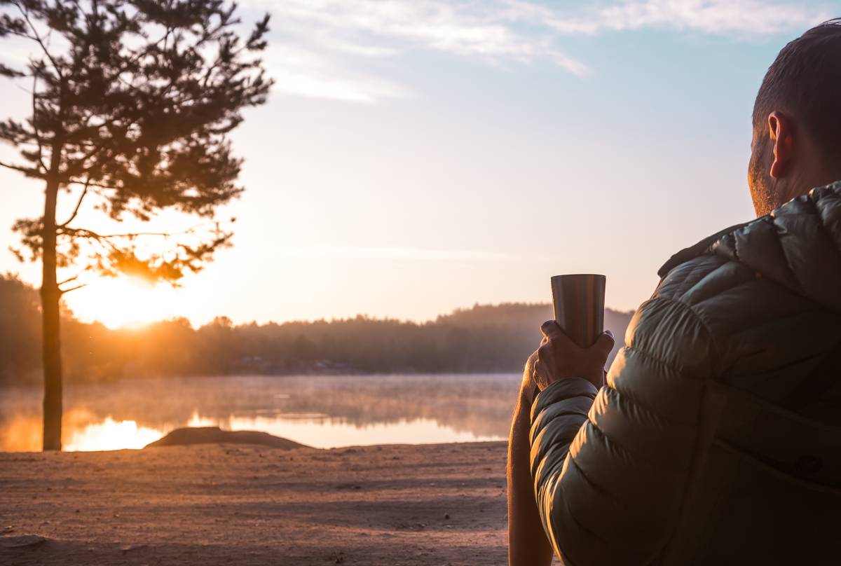 Man drinking a coffee from a thermos watching the sunrise over a lake