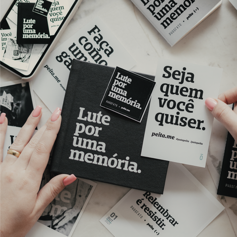 foto colorida em ambiente interno de uma mão branca com unhas cor salmão segurando um álbum preto com a frase lute por uma memória na esquerda e na direita uma foto de um cartaz escrito seja quem você quiser. Tem também um celular com foto do envelope preto, quadrado escrito lute por uma memória. 