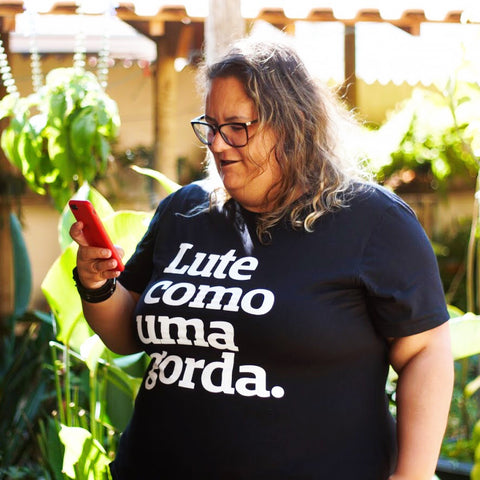 foto colorida da jessica, mulher branca, cabelos castanhos claros, médios, lisos, vestida com uma camiseta preta com a frase lute como uma gorda em branco. Está olhando o celular. 