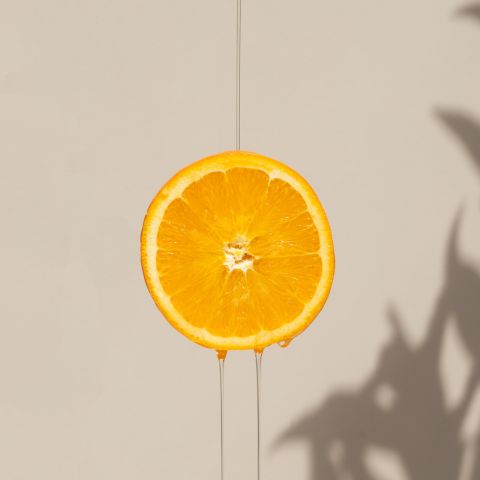 A fresh orange slice glistens with droplets of oil as it rests on a smooth beige background. The vibrant orange color of the slice contrasts beautifully with the neutral backdrop, while the glistening oil adds a touch of freshness and juiciness to the image.