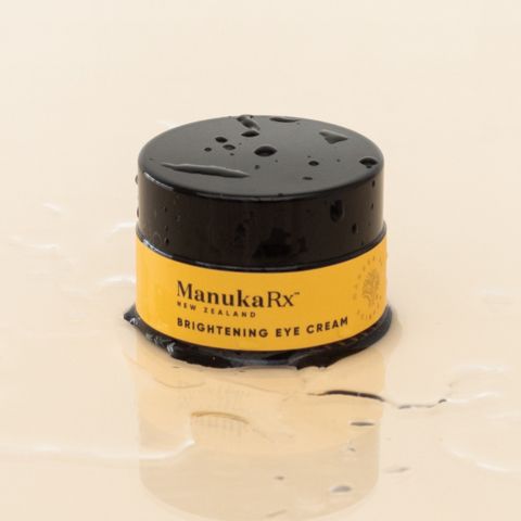 Pot of Brightening Eye Cream on a beige background. The pot has a light orange label with the product name. Drops of water can be seen around the pot, adding a refreshing touch
