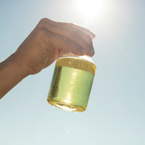 Mānuka Essential Oil and Rosehip Oil: A Collagen Boosting Duo image showing a bottle of pure East Cape Manuka oil glowing golden in the sun held up by a male's hand against a blue sky.