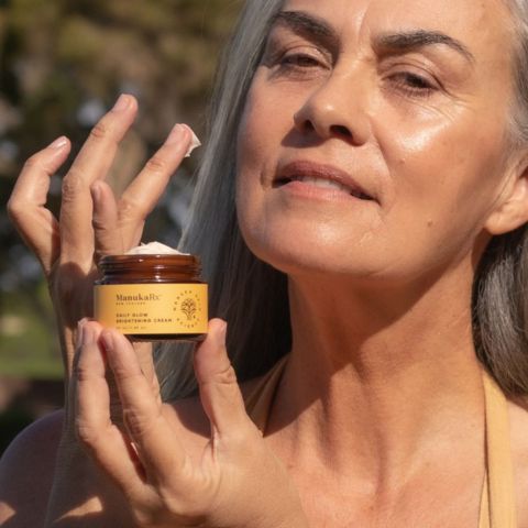 Graceful woman with stunning grey hair donning a vibrant yellow dress, showcasing radiant skin while delicately holding a jar of Daily Glow Brightening Cream, enhancing her natural luminosity.