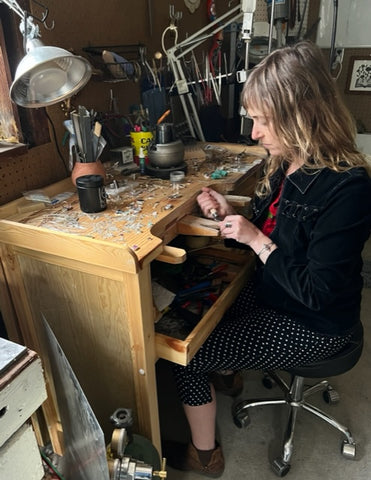 artist wearing black at a dimly let jewelry bench, setting a stone with a bezel rocker tool. She has long, messy strawberry blonde hair.