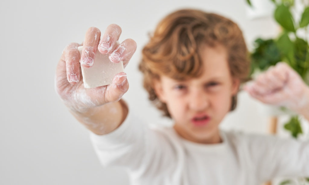 An enthusiastic boy with a mischievous grin holds up a sudsy hand, proudly displaying a bar of handmade soap.