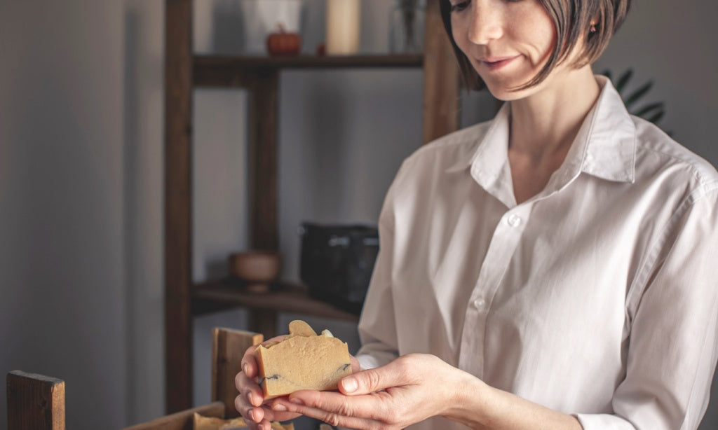 An accomplished female soap maker smiles warmly as she gazes down at a freshly crafted bar of soap, radiating pride and satisfaction in her creation.