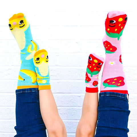 child and adult feet in the air, each wearing Pals Socks: banana socks on one foot and strawberry socks on the other